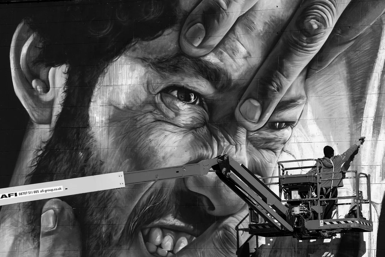 Liam Bononi portfolio photograph: a large monochrome mural of a face framed by hands, with a painter on a raised platform.